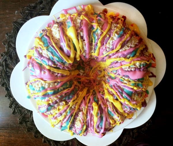 Easy Rice Krispies Cake top view of a round bundt rice krispies cake with different colored bright chocolate drizzled over it on a white plate shaped like a flower