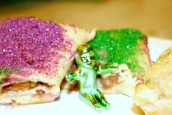 pieces of king cake with purple sugar icing and green purple icing with a tiny green plastic King Cake baby sitting between the two pieces of King Cake