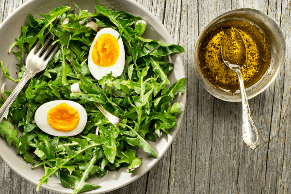 Dandelion salad with boiled egg in a bowl on a table with oil dressing in a cup next to it