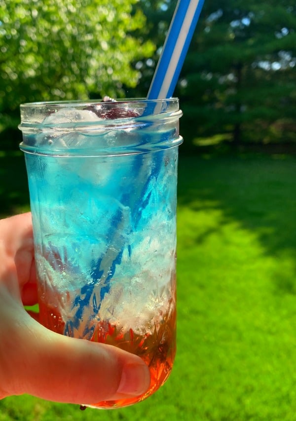 How to Make Layer Drinks glass held up to the sun with blue white and red layered drink