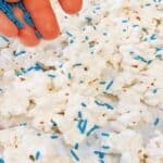 The best white chocolate popcorn recipe spread on a single layer on parchment paper baking sheet with a hand putting blue sprinkles on the white coated popcorn