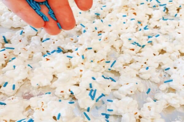 The best white chocolate popcorn recipe spread on a single layer on parchment paper baking sheet with a hand putting blue sprinkles on the white coated popcorn