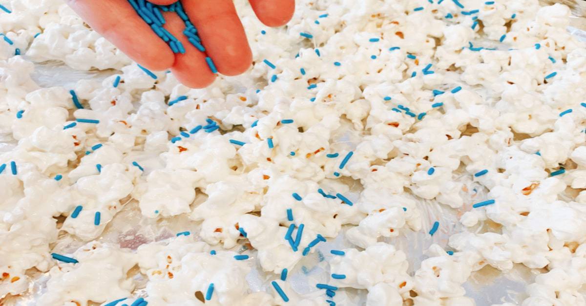 The best white chocolate popcorn recipe spread on a single layer on parchment paper baking sheet with a hand putting blue sprinkles on the white coated popcorn