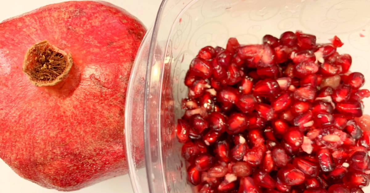 how to cut open pomegranate with a whole pomegranate sitting next to pomegranate arils seeds in a clear bowl