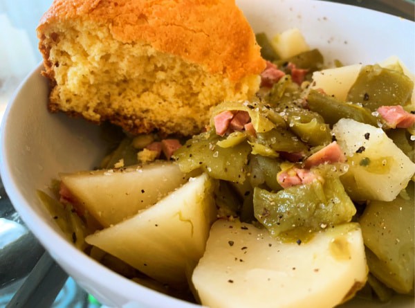 green beans and potatoes in a white bowl with a piece of cornbread sitting on the side