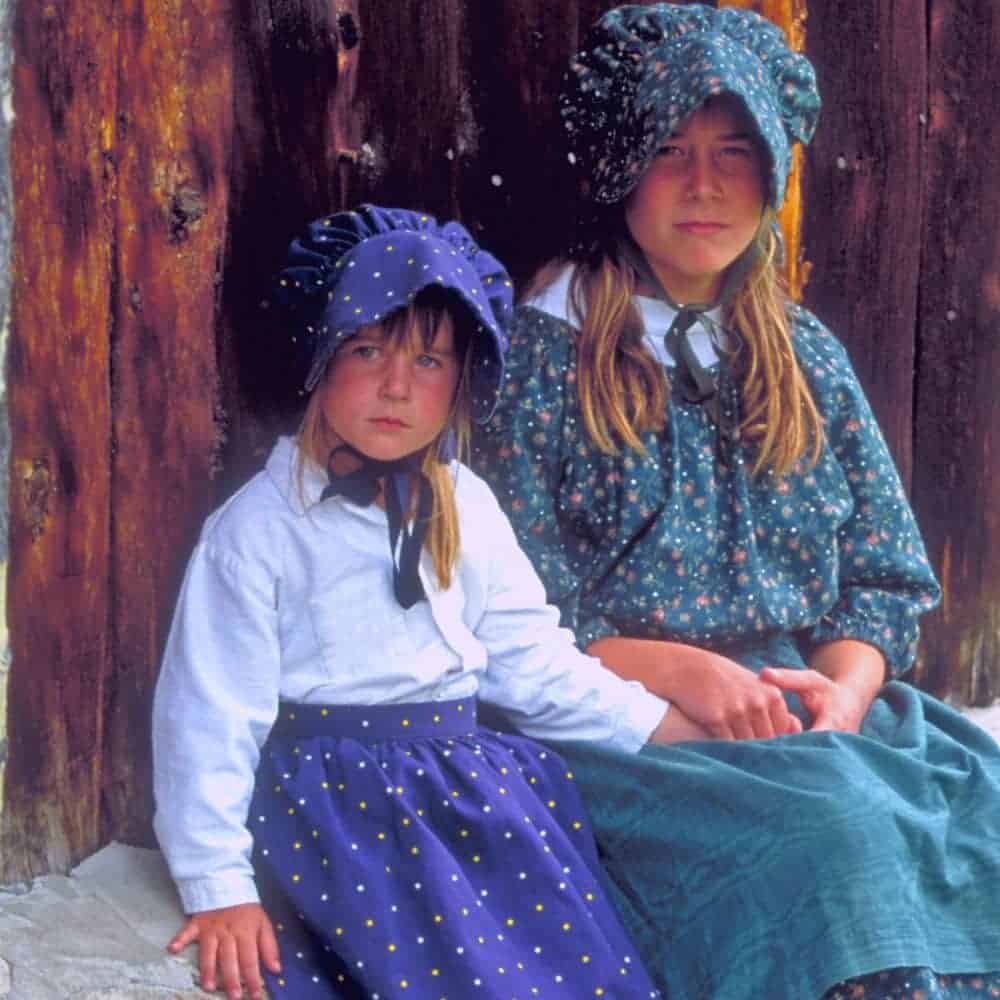 Pioneer Party Costumes For Kids two girls dressed in prairie dresses and bonnets sitting on a porch