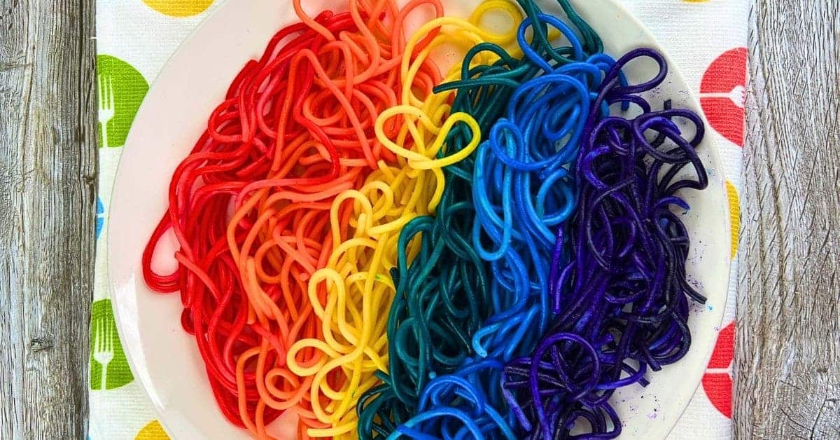 Colored Spaghetti Recipe For Kids (How To Make Rainbow Noodles) on a white plate and a dishtowel with colored forks