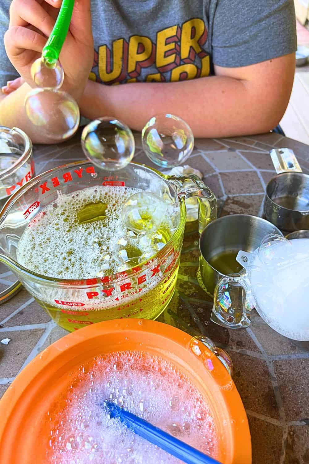 DIY Bubble Wand Solution (best bubble solution recipe) hands of a child blowing bubbles homemade through a straw with multiple big bubbles floating in the air