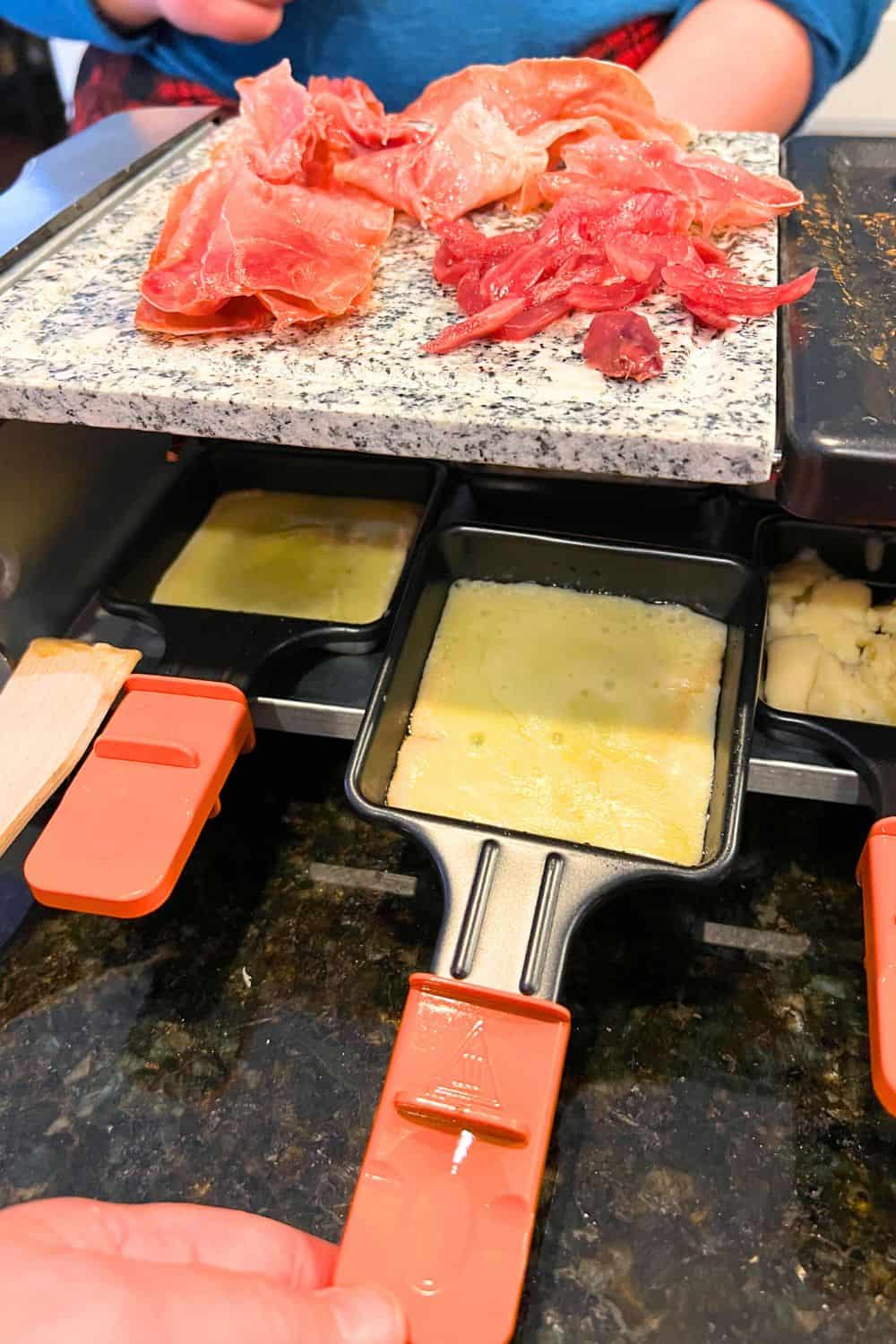 What is a raclette maker - home raclette grill on counter with prosciutto on grill top and melting raclette in trays under the grill
