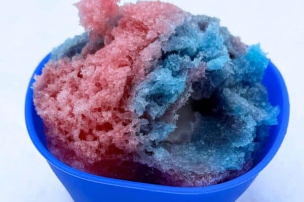 Homemade Snow Cones Recipe With Snow Cream - Shave Ice Syrup Recipe / and Shaved Ice Recipe Snow Cones Recipes sitting in a blue snow cone cup in the snow with red and blue Sno Cone Syrup