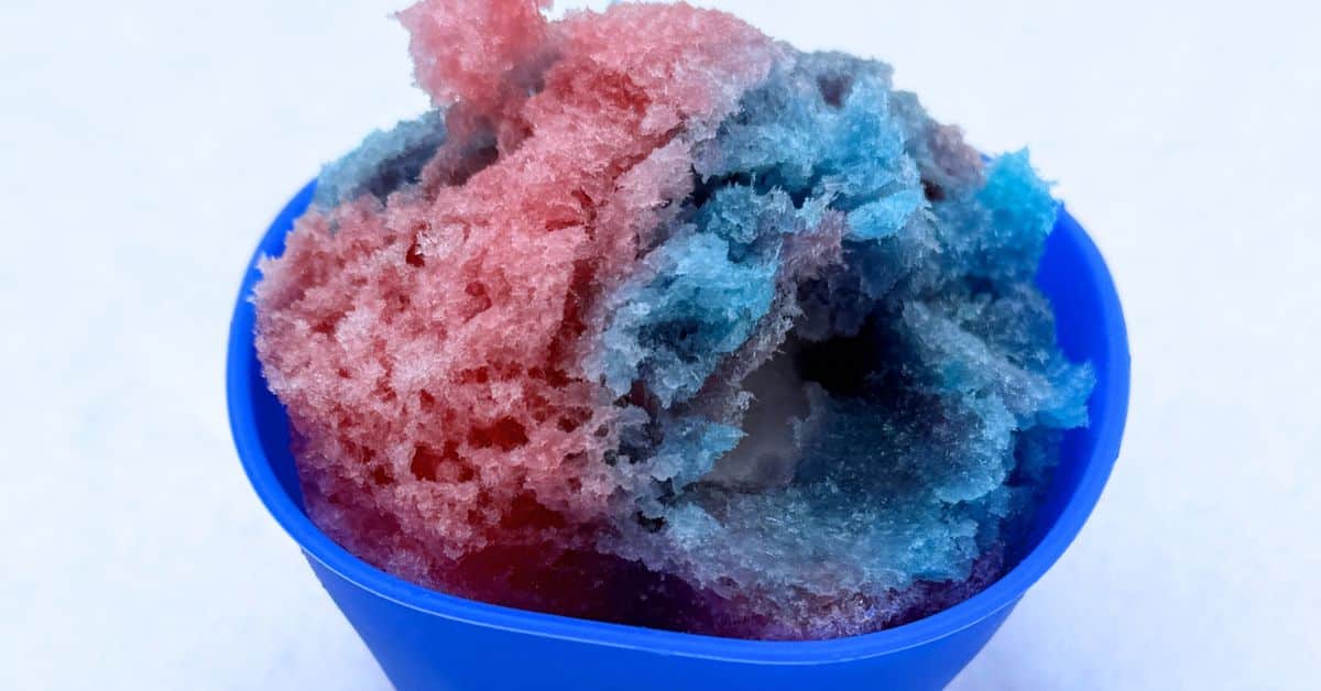 Homemade Snow Cones Recipe With Snow Cream - Shave Ice Syrup Recipe / and Shaved Ice Recipe Snow Cones Recipes sitting in a blue snow cone cup in the snow with red and blue Sno Cone Syrup